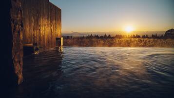 A wooden building with a pool extending from its side, set against a backdrop of a sunset over a forested landscape.