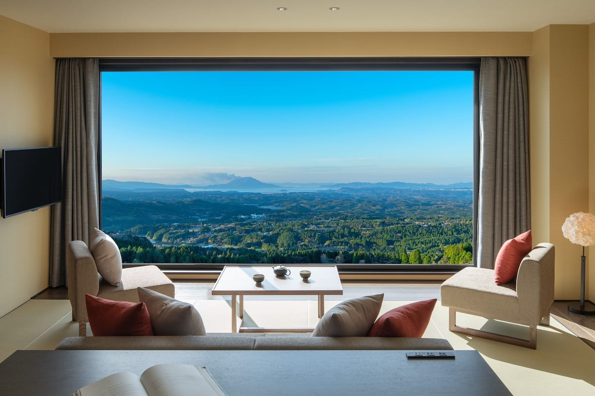 A modern hotel room with a large window offering a scenic view of a valley and mountains. There is a sofa, a small table with a tea set, and a flat-screen TV mounted on the wall.