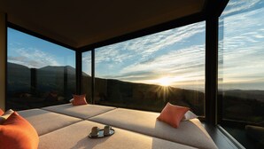A modern room with large windows offering a view of a mountainous landscape at sunset.