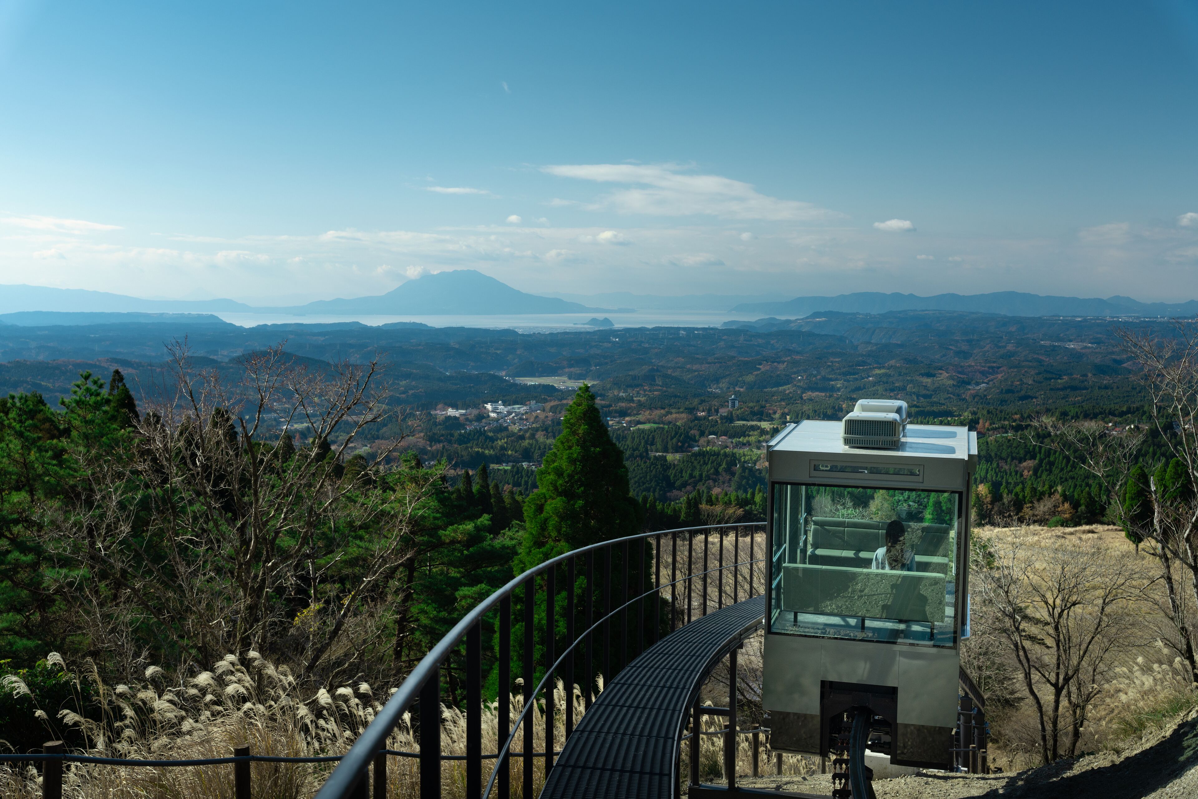 A glass observation tower with a metal railing, offering panoramic views of a mountainous landscape and a body of water.