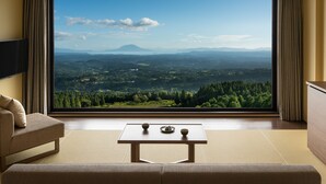 A room with a large window offering a view of a mountainous landscape. There is a sofa, a small table with two bowls, and a flat-screen TV mounted on the wall.