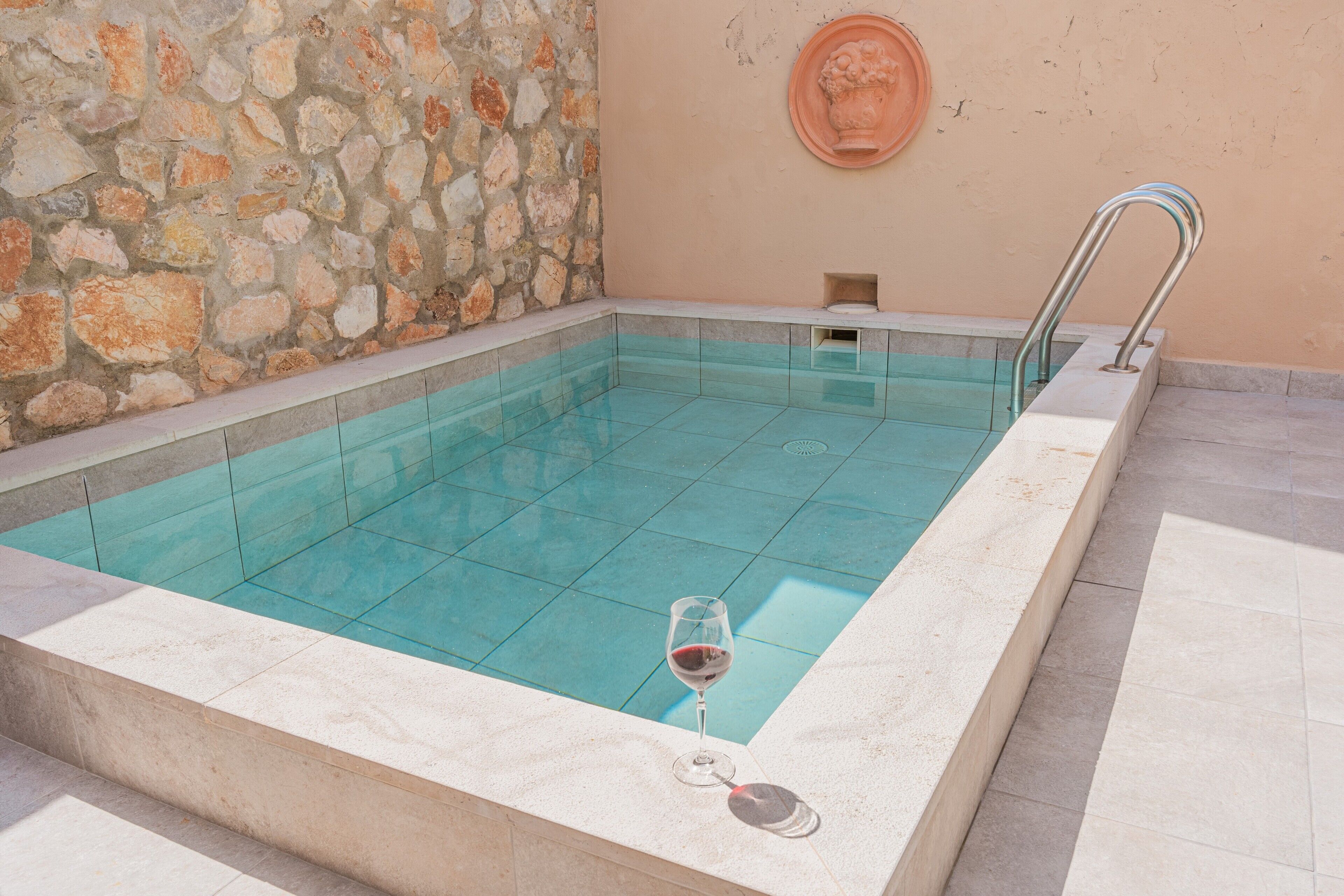 Una piscina rectangular con agua azul cristalina, una pared de piedra y una escalera metálica.