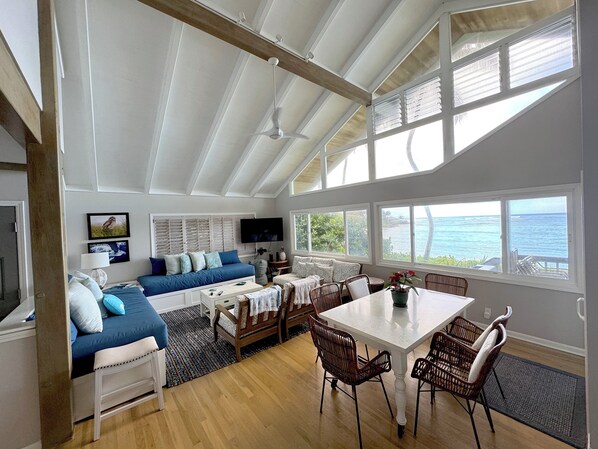 Ein geräumiger Wohnbereich mit großem Fenster zum Meer, ein Essbereich mit Korbstühlen und eine gemütliche Sitzecke mit blauem Sofa und gemusterten Kissen.