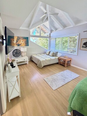 A bedroom with a bed, a desk, a chair, a standing fan, and a window with blinds.