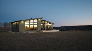 Une maison moderne, de plain-pied, dotée de grandes fenêtres et d’un toit plat, située dans un champ verdoyant, avec un paysage vallonné en arrière-plan.