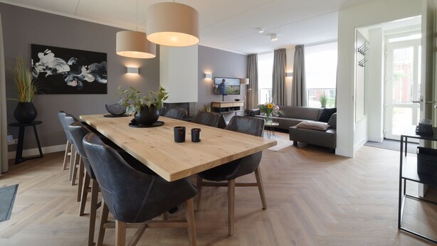 En moderne stue med et træspisebord, sorte læderstole, en grå sofa og et fjernsyn monteret på væggen.