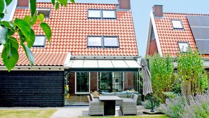 En boligbygning med rødt tegltag, en overdækket terrasse med havemøbler, og en have med forskellige planter.