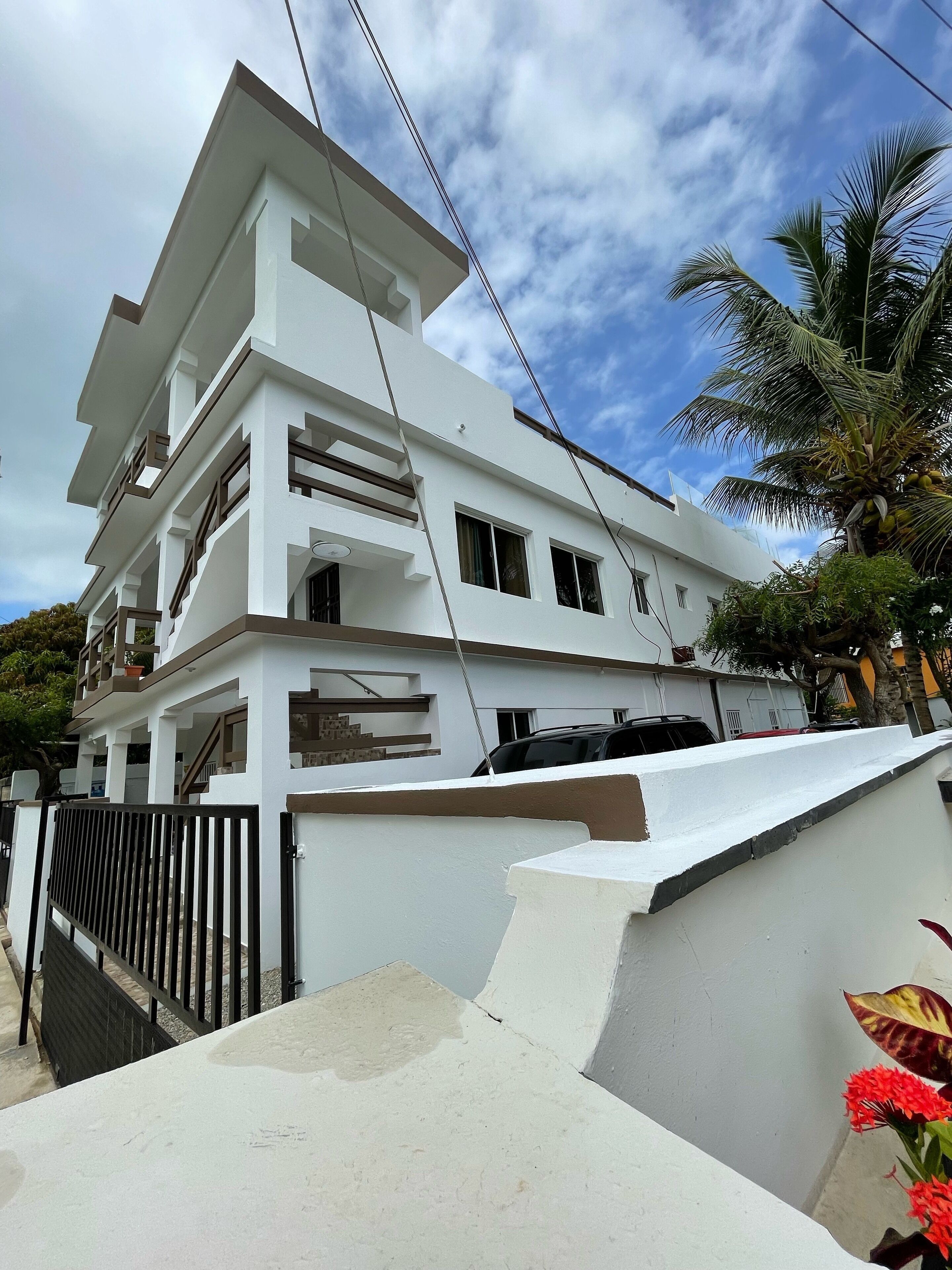 A multi-story white building with balconies, a gated entrance, and a palm tree in the foreground.