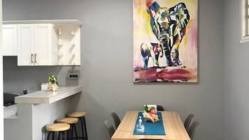 Una cocina moderna con armarios blancos, una mesa de comedor de madera con sillas negras y un cuadro abstracto y colorido en la pared.