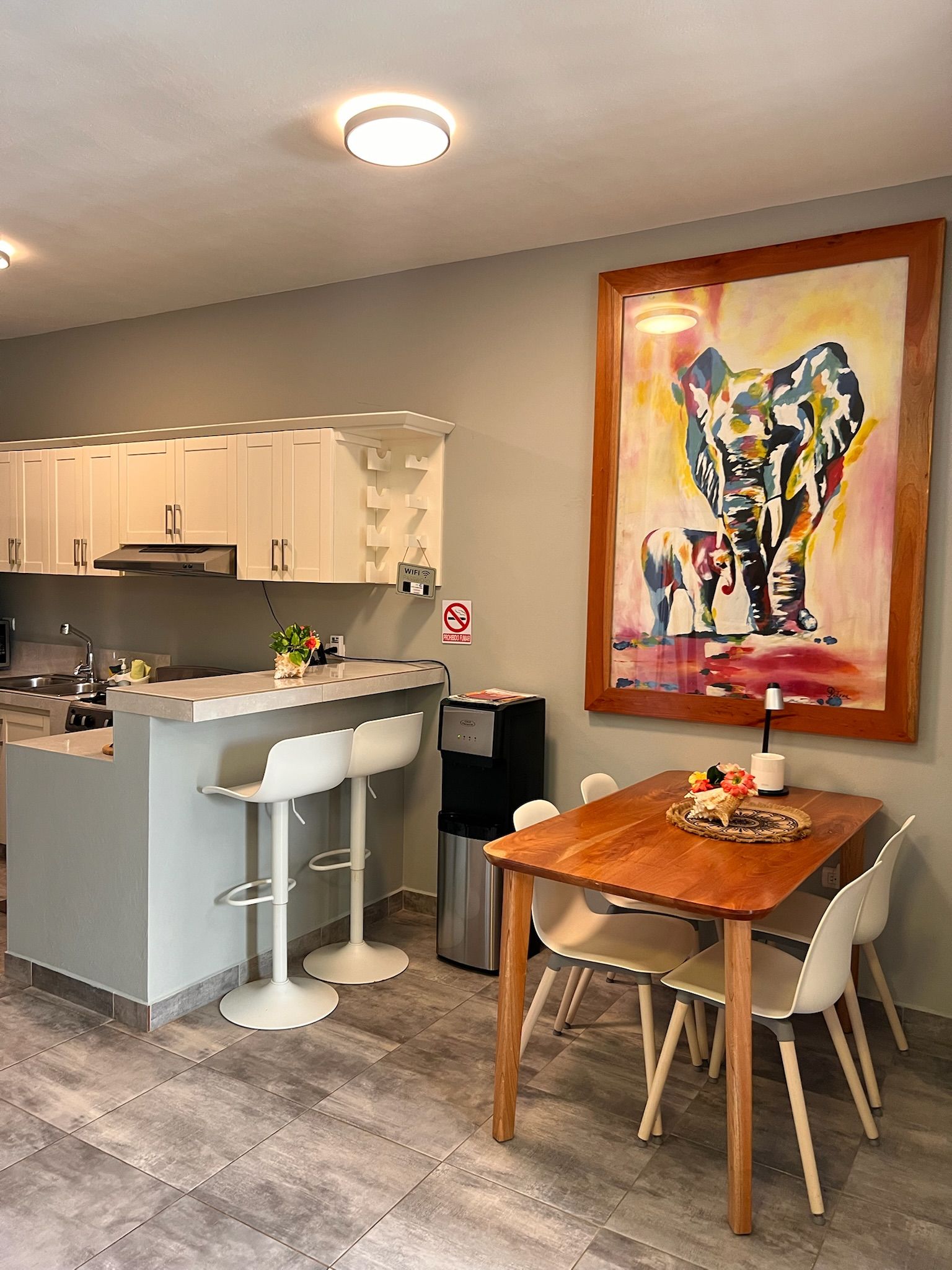Una cocina moderna con armarios blancos, una isla central con taburetes, una mesa de comedor con sillas y un cuadro abstracto grande en la pared.