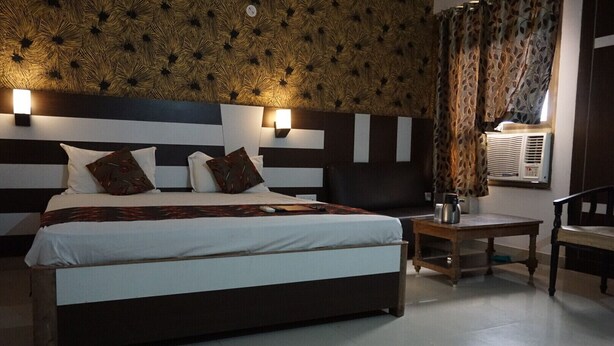 A hotel room with a large bed, two bedside tables, a chair, a wall-mounted heater, and a patterned wall. - Hotel Mani International (Patna)