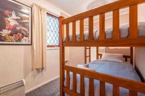 A bunk bed with a wooden frame, a single bed with a grey blanket, a window with a diamond pattern, and a framed floral painting on the wall.