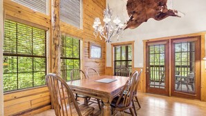 Un comedor con mesa y sillas de madera, ventanas grandes con persianas y una lámpara de araña.