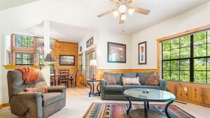A cozy living room with a sofa, armchair, and coffee table, wooden walls, large windows, and ceiling fan. - Roomy 2BR+Loft Walk-in Cabin near Shows, Shopping, and Dining! (Branson)