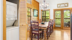 A dining area with a wooden table and chairs, a chandelier, and a view through large windows. - Roomy 2BR+Loft Walk-in Cabin near Shows, Shopping, and Dining! (Branson)