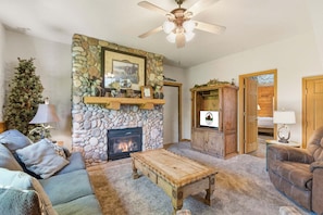 A cozy living room with a stone fireplace, a ceiling fan, and a television.