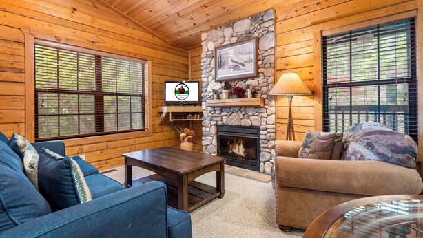 A cozy cabin interior with a stone fireplace, wooden walls, a blue sofa, and a television. - Romantic Walk in Cabin with King Bed, Fireplace & Whirlpool Tub (Branson)