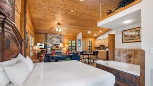 A spacious bedroom with a large bed, a sitting area with a table and chairs, a kitchen in the background, and a bathroom with a bathtub. - Romantic Walk in Cabin with King Bed, Fireplace & Whirlpool Tub (Branson)
