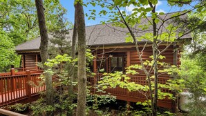 A rustic cabin surrounded by dense forest, with a wooden deck and a large window. - Romantic Walk in Cabin with King Bed, Fireplace & Whirlpool Tub (Branson)