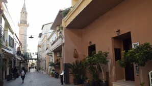 Une ruelle étroite pavée, bordée d’immeubles, dont certains sont dotés de balcons et de plantes en pot.