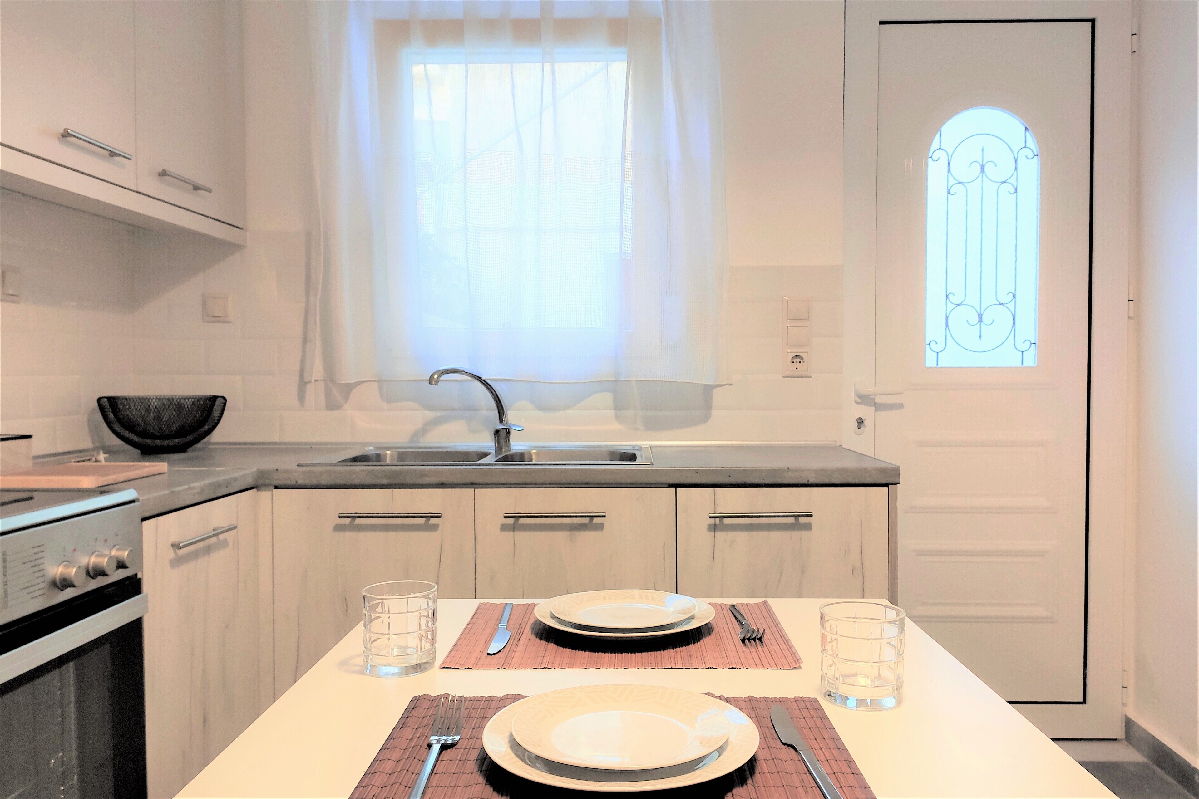A modern kitchen with a dining area set for two, featuring a sink, stove, and a window with a decorative glass door.