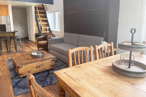 Un espacio habitable compacto con sofá, mesa de comedor de madera y sillas. Una escalera conduce a un nivel superior.
