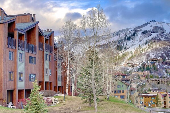 A multi-story wooden building with balconies, a ski resort in the background, and a mountainous landscape.