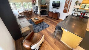 A modern living room with a stone fireplace, a flat-screen TV mounted above it, a dining area with a wooden table and chairs, and a kitchen area with a black countertop and bar stools. - Steps to Mtn/Resort/Free Town Bus (Across St)+ Heated Pool-HotTub-Grill-Updated-Spacious-2 Levels (Steamboat Springs)