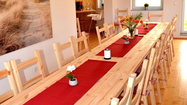 Une longue table à manger en bois, agrémentée de chemins de table rouges, des chaises en bois avec des coussins à carreaux, et un tableau représentant une plage accroché au mur.