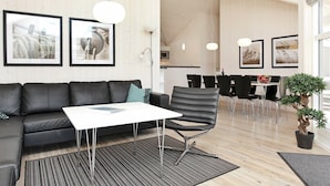 A modern living room with a black leather sofa, a white coffee table, and a black office chair. There are framed pictures on the wall, a potted plant, and a dining area in the background. - Holiday Home in Großenbrode (Grossenbrode)