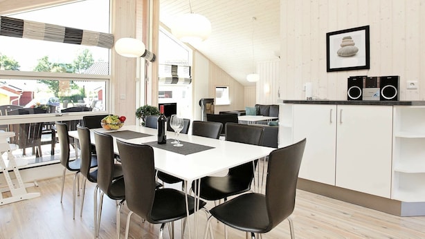 A modern kitchen with white cabinets, a dining area with a white table and black chairs, and a large window providing a view of the outside. - Holiday Home in Großenbrode (Grossenbrode)