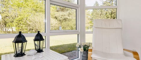 Una habitación con ventanas amplias, una silla blanca y dos faroles negros sobre una mesa blanca.