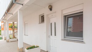 Un bâtiment blanc avec une grande fenêtre, une porte et une petite fenêtre au-dessus de la porte. Un jardinier blanc rempli de fleurs se trouve à côté de la porte.
