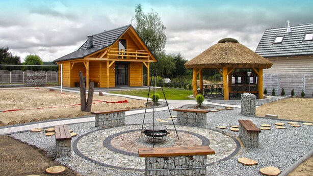 A wooden cabin with a thatched-roof gazebo, surrounded by a gravel courtyard with benches and a fire pit. - Eco-friendly House by Goszcza Lake (Lubrza)