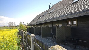 A wooden building with a sloped roof, a fenced area with a bench, and a field of yellow flowers. - Cozy Studio 2 at the Seaside Agrotourism (Darlowo)