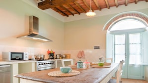 Una cocina con una mesa de madera rústica, una estufa y un microondas. Hay una ventana con cortinas y una puerta con marco verde claro.