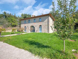 A stone building with a red-tiled roof, a central arched doorway, and a small garden with a tree and shrubs. - Villa in Tuscany With Private Pool (Castiglion Fiorentino)