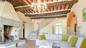 Un amplio salón con un sofá blanco, cojines verdes, una chimenea, una escalera y una araña.