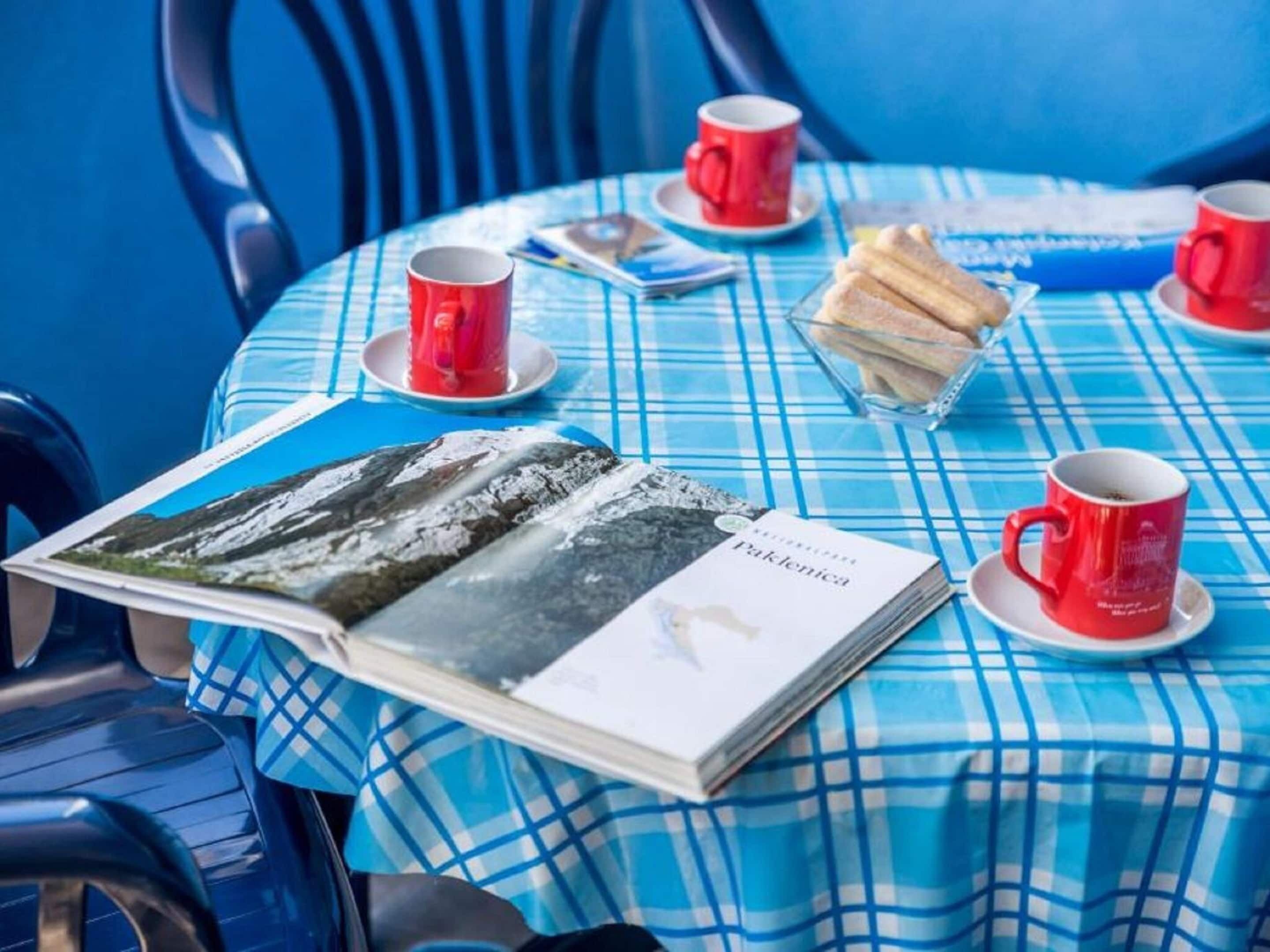 A table with a blue checkered tablecloth, a magazine, red cups, and a basket of bread.