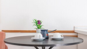 Uma mesa redonda pequena com duas chávenas e pires brancos, uma chaleira azul e uma planta em vaso com folhas verdes e uma flor roxa.