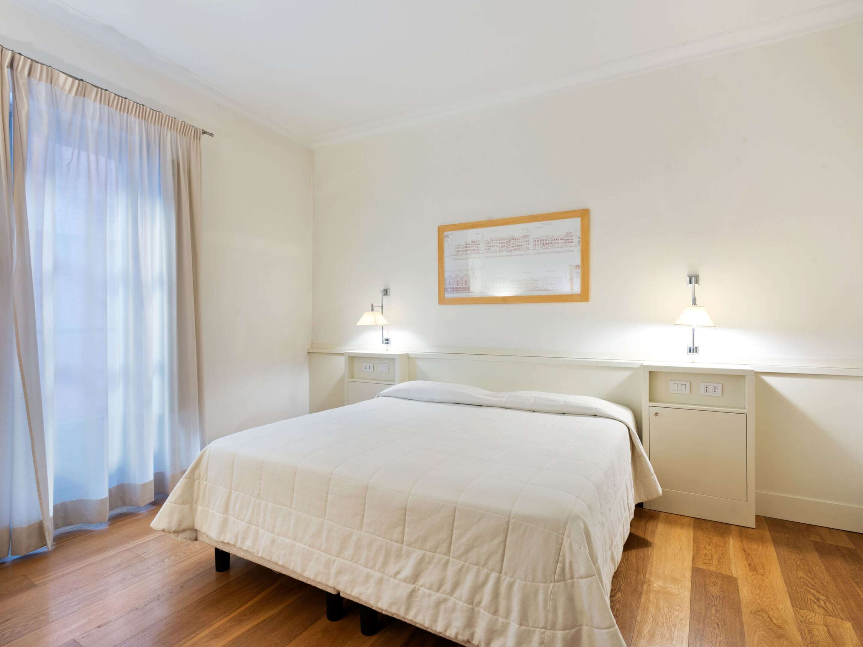A hotel room with a bed, wooden flooring, a window with curtains, and two bedside lamps.