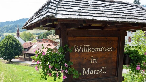 A wooden sign with the text "Willkommen In Marzell" surrounded by greenery and a rural landscape.