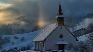 A church with a steeple and a cross, surrounded by snow-covered landscape and trees.