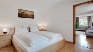 A neatly made bed in a room with a wooden floor, a painting on the wall, and a view of another room through an open door.