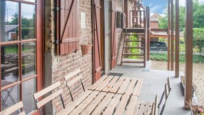 A covered outdoor seating area with wooden benches, a brick wall, and a view of a garden and buildings in the background. - Splendid Villa in Vorsen (Gingelom)