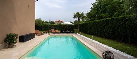 A rectangular swimming pool with lounge chairs and towels, surrounded by a landscaped garden and a hedge.
