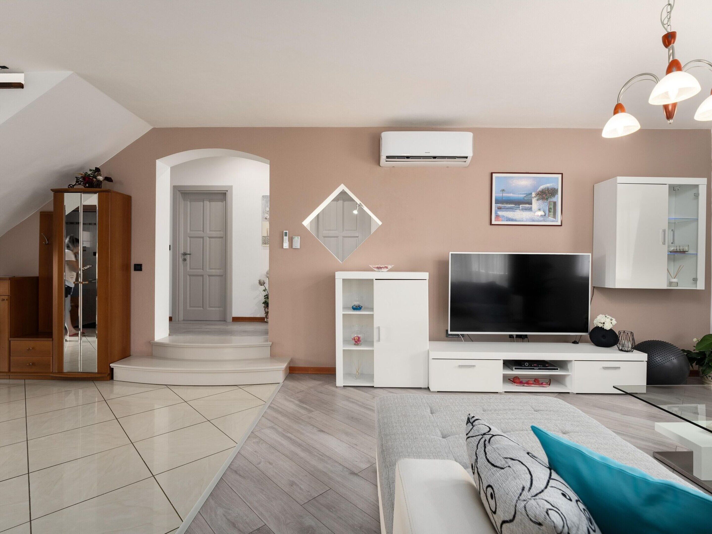 A modern living room with a flat-screen TV, white cabinetry, a mirror, and a staircase leading to an upper level.