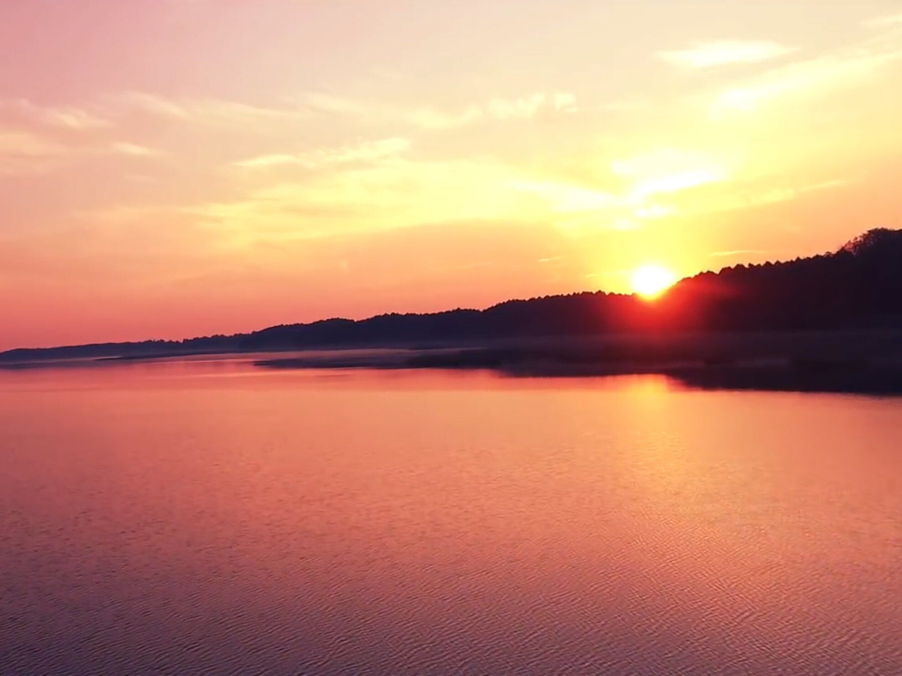 Ein ruhiger See bei Sonnenuntergang, im Hintergrund eine Hügelsilhouette.