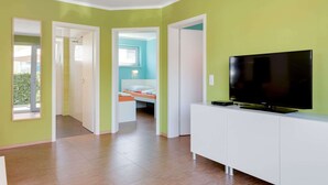 Una habitación moderna con una televisión grande sobre un mueble blanco, una cama al fondo y una puerta que da a un balcón.
