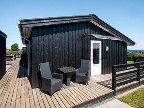 A wooden deck with two wicker chairs and a small table, a black building with a white door, and a view of the sea.